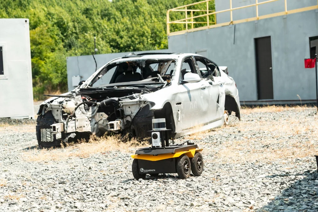 A yellow wheeled robot with mounted sensors on gravel in front of a damaged car at the ArtIAMAS 2024 test site