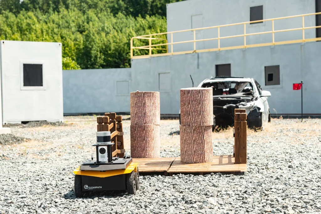 A wheeled robot beside a wooden platform with cylindrical obstacles in the outdoor test area