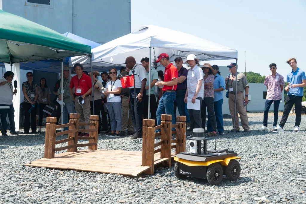 Participants watching a wheeled robot beside a wooden platform during the ArtIAMAS 2024 field demonstration