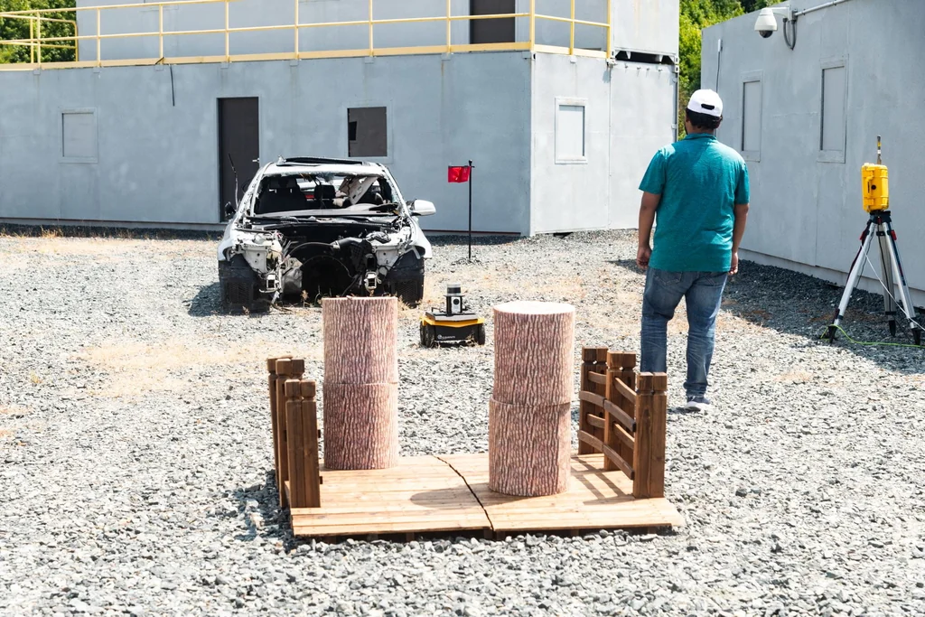 A participant beside a wheeled robot and a wooden obstacle course in the ArtIAMAS 2024 test area