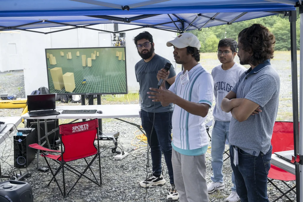 Participants presenting a robotics experiment beside a simulation display under a blue canopy at ArtIAMAS 2025