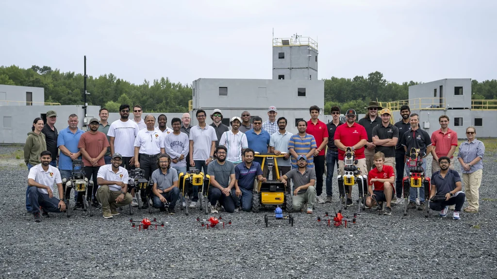 ArtIAMAS 2025 participants posing outdoors with quadruped robots, wheeled robots, and drones