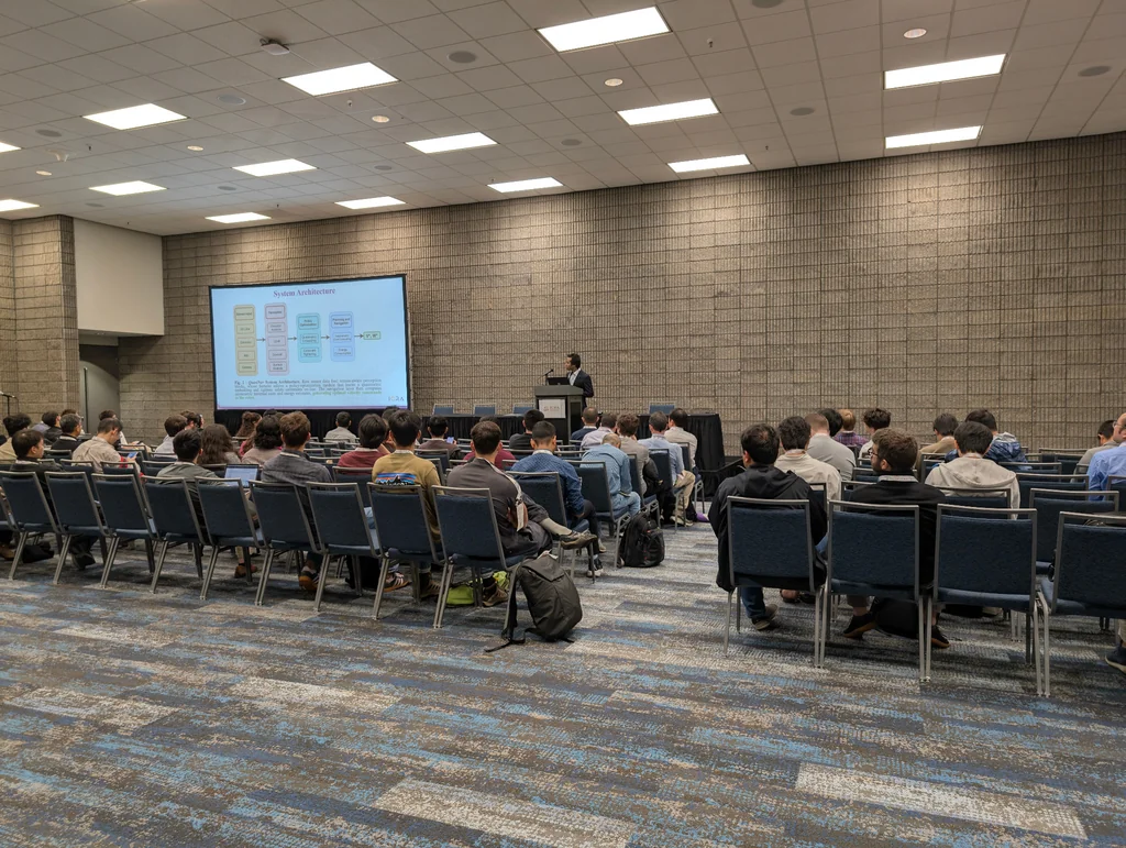 A speaker at the podium beside a projected system architecture slide, with the audience seated in the ICRA 2025 presentation room